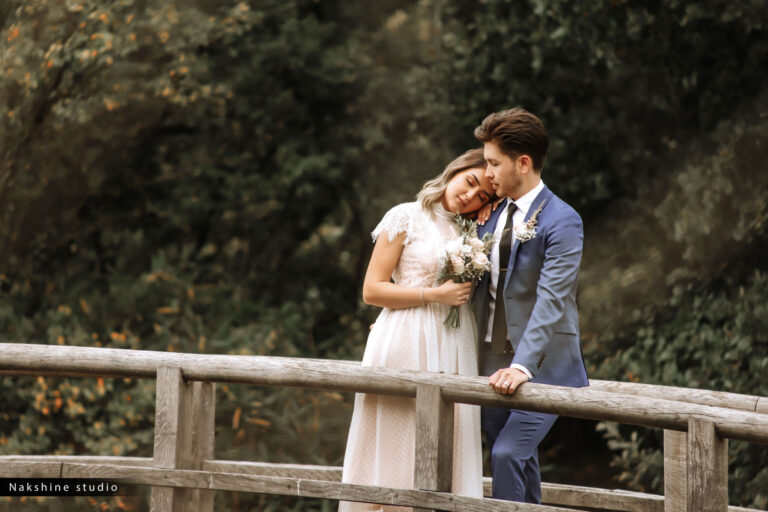 Een bruidspaar omhelst elkaar liefdevol op een houten brug in een natuurlijke omgeving.