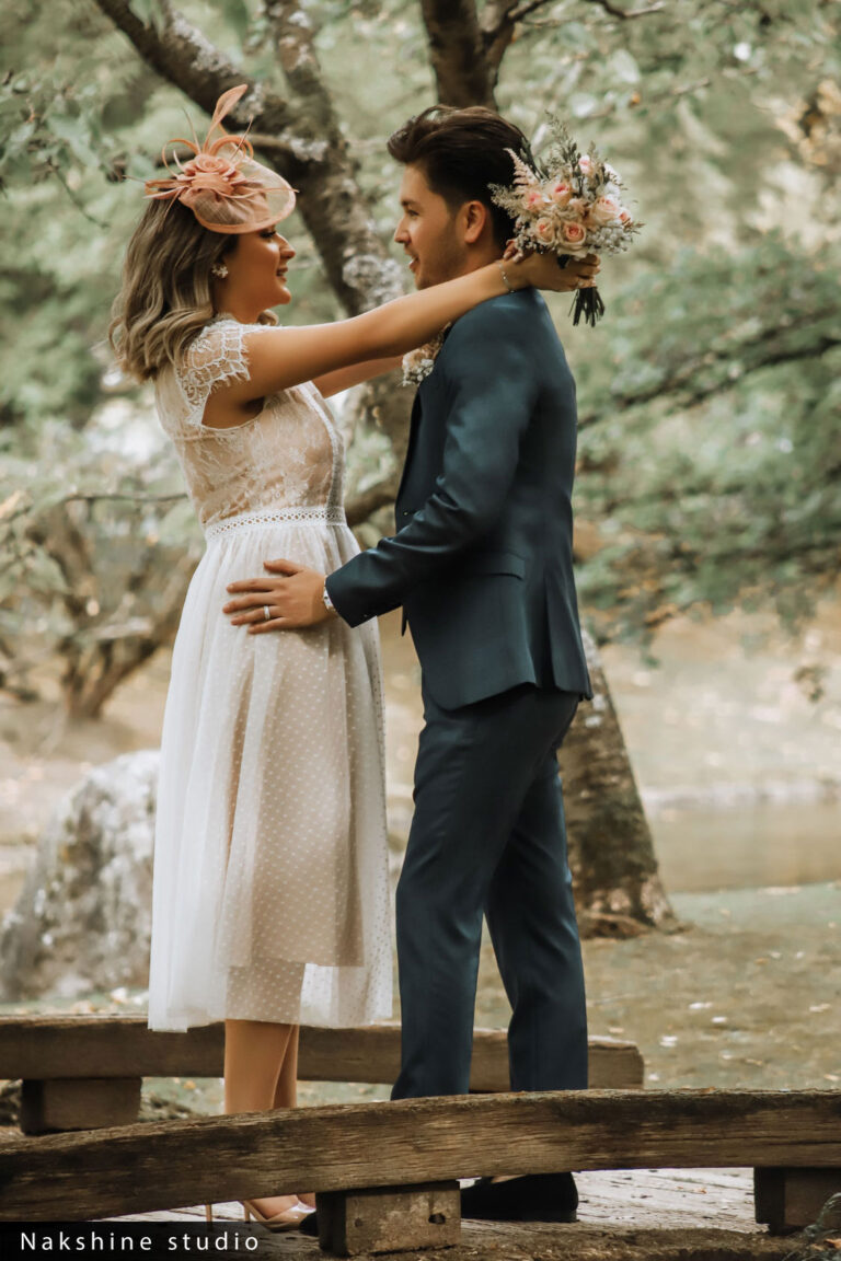 Een bruidspaar staat liefdevol in een bosrijke omgeving, de vrouw draagt een korte trouwjurk en een bloemenhoofdtooi, de man in een blauw pak.